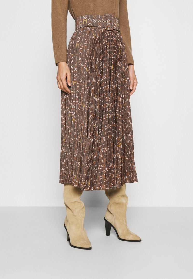 Femme portant une jupe midi plissée à motif marron avec une ceinture assortie et des bottines à talons en daim beige, debout devant un fond uni.