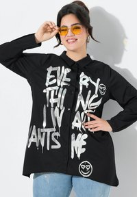 Zwarte overhemdd met zilver graffiti-achtige tekst, featuring een smiley face graphic. Lange mouwen en een ontspannen pasvorm. Draagt over lichtblauwe jeans.