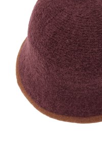 Bordowy filcowy kapelusz o zaokrąglonym kształcie i kontrastowym brązowym rondzie. Faktura jest miękka, z delikatnie meszkiem widocznym na powierzchni.