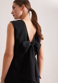 Robe noire avec un profond décolleté en V dans le dos. Un grand nœud à l'arrière apporte un intérêt visuel, dans une texture de tissu lisse.