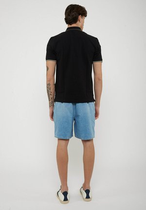 Uomo in piedi di spalle, indossa una polo nera a maniche corte, pantaloncini di jeans azzurri, scarpe da ginnastica bianche, con un tatuaggio sull'avambraccio sinistro.