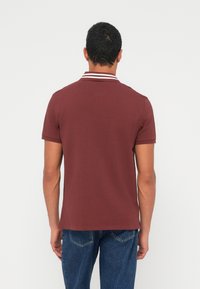 Egy férfi háttal, rövid ujjú, csíkos galléros bordó galléros pólót és kék farmert visel, egyszínű fehér háttér előtt.