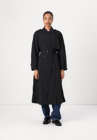 Trench coat nero con bottoni doppi, vita con cinturino, polsini con bottoni e design di lunghezza media. Indossato sopra jeans blu e scarpe.