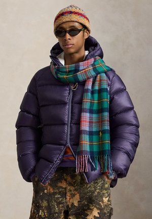 Personne portant une doudoune violette, un bonnet tricoté coloré, une écharpe à carreaux, des lunettes de soleil et un pantalon camouflage, debout les mains dans les poches.