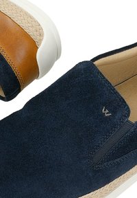 Chaussure à enfiler en daim bleu marine avec un accent en cuir marron au talon, semelle en caoutchouc blanche et petit logo de marque en métal sur le côté.