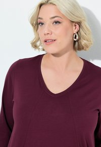 Bordeaux lange mouw top met een V-hals en naaddetails, gemaakt van zachte stof. Het model heeft kort, golvend blond haar.