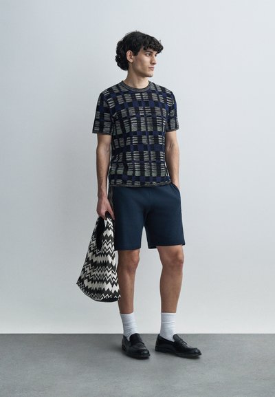 Πουκάμισο με κοντά μανίκια σε ναυτικό μπλε χρώμα, με συνδυασμό ρίγες και καρό. Συνδυασμένο με σκούρες βερμούδες και μια τσάντα tote μαύρου και άσπρου με ζιγκ-ζαγκ σχέδιο.