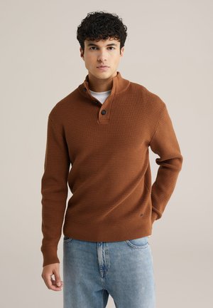 Brun ribbet sweater med høj krave, knapdetaljer og struktureret stof. Båret med lyseblå jeans, der har en afslappet pasform.
