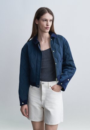 Γυναίκα που φοράει μπλε σκούρο μπουφάν με καπιτονέ υφή πάνω από σκούρο γκρι αμάνικο τοπ και λευκό σορτς με ψηλή μέση, στέκεται με το ένα χέρι στην τσέπη.