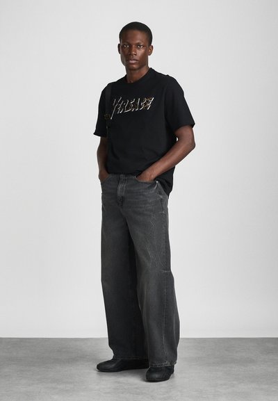 T-shirt preta com lettering metálico, combinada com calças largas cinzas. O sujeito usa sapatos pretos, de pé com as mãos nos bolsos contra um fundo neutro.