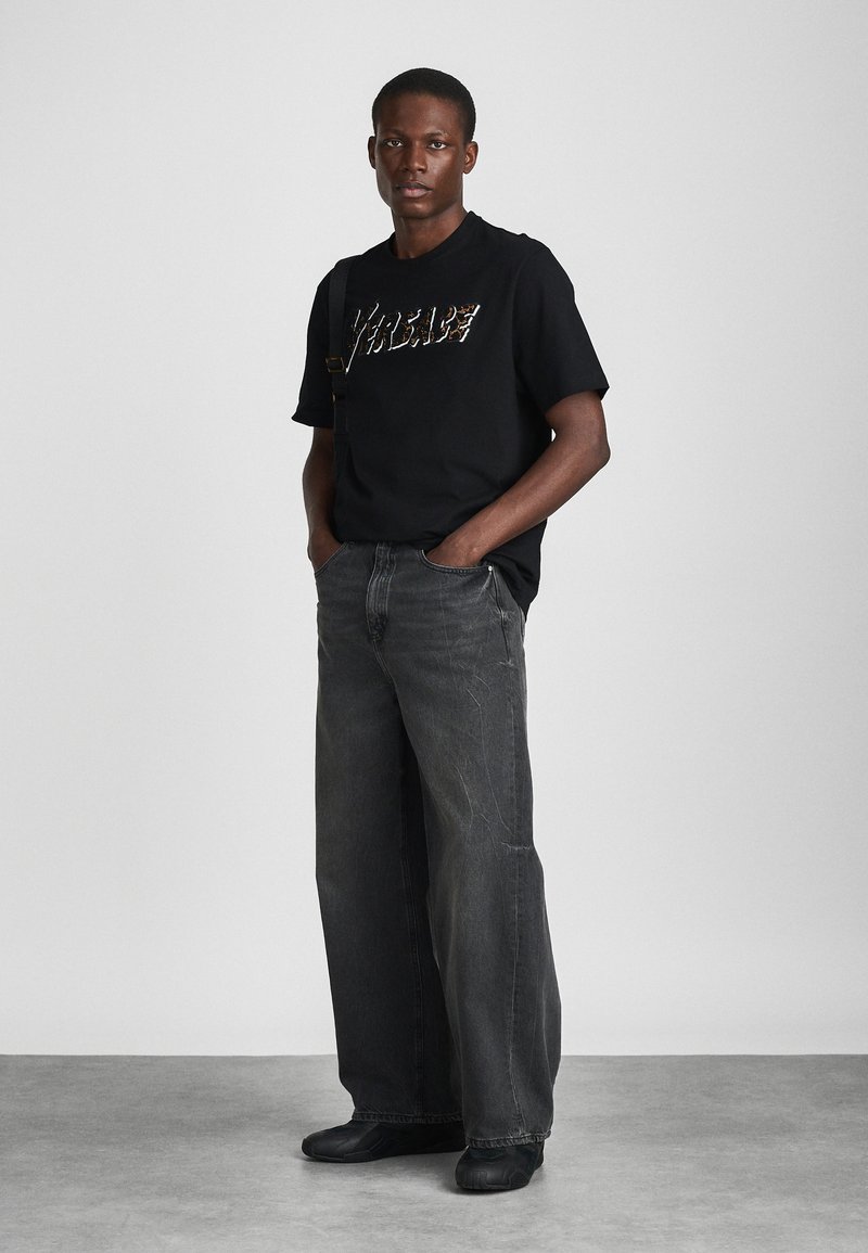 T-shirt noir avec des lettres métalliques, associé à un pantalon large gris. Sujet portant des chaussures noires, debout avec les mains dans les poches devant un fond neutre.