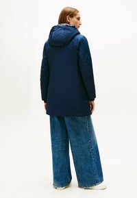 Giacca imbottita blu navy con cappuccio, caratterizzata da una texture liscia e da un taglio più lungo. Abbinata a jeans in denim sbiaditi a gamba larga.