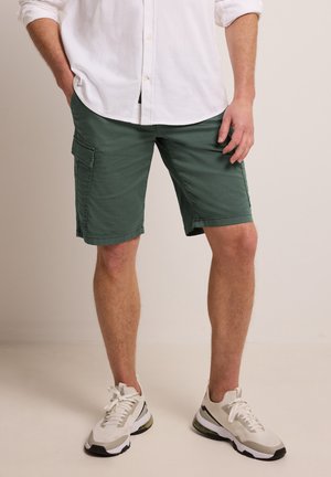 Mann trägt grüne Cargo-Shorts, weißes, hochgekrempeltes Hemd und weiße Turnschuhe, steht vor einem schlichten Hintergrund.