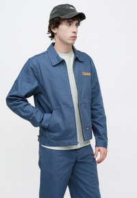 Μπλε τζάκετ από καμβά με γιακά που στέκεται και φερμουάρ μπροστά, με πλαϊνές τσέπες και κεντημένο λογότυπο σε πορτοκαλί. Φοράται πάνω από γκρι μπλουζάκι.