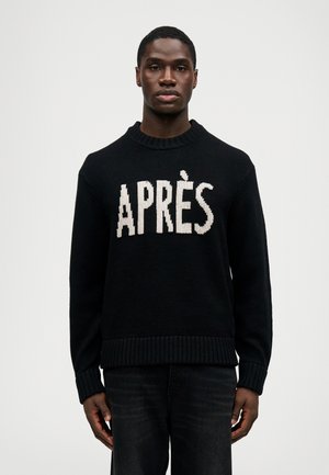 Melna adīta džempera, kurā uzraksts "APRÈS" balta krāsā, ar apaļu apakšējo daļu un ribotām aprocēm un apakšmalu. Apvienots ar tumšām džinsām.