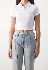 Kvinna i en vit kortärmad polo-tröja och högmidjade ljusblå cargobyxor med knäppta fickor.