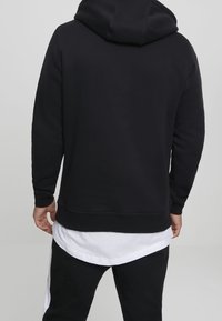 Svart hoodie av mjukt tyg med avslappnad passform, med en känguruficka och en stor huva. Vit skjortslut synligt under.