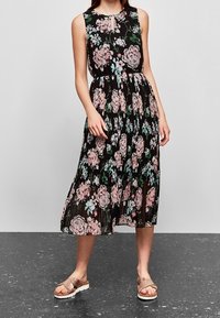 Schwarz-blumiges Kleid mit rosa und grünen Rosenmustern, ärmelloses Design, plissierter Rock und ein Schlüssel-Ausschnitt am Hals. Getragen mit braunen Sandalen.