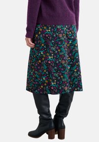 Een rok met bloemenmotief in donkere stof, met levendige groene, paarse en gele tinten. Gepaald met zwarte kniehoge laarzen en een paarse trui.