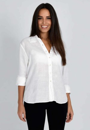 Donna sorridente con lunghi capelli scuri che indossa una camicia bianca con bottoni, maniche arrotolate e pantaloni neri su uno sfondo semplice.