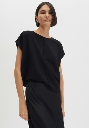 Giovane donna con capelli corti che indossa un top nero largo e una gonna, in piedi davanti a uno sfondo chiaro e uniforme.