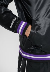 Chaqueta bomber de satén negra con puños acanalados en rayas moradas y blancas y un logotipo bordado en la manga.