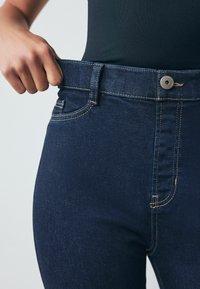 Tumed sinine denim tasku püksid, kõrge vöökohaga, kontrastsete õmblustega ja nupuga sulgemine. Nähtav esipocket detail. Sile pind.