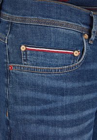 Denim jeans featuring a dark blue wash, gold hardware, a back pocket with red, white, and blue striped detail, and subtle texture.