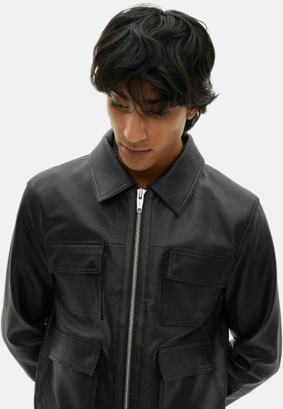 Young man with dark wavy hair wearing a black leather jacket with four front pockets and a silver zipper, looking downward.