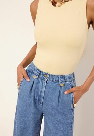 Hihaton beige toppi yhdistetty korkeavyötäröisiin, laskostettuihin sinisiin farkkuihin, asusteena massiivinen helminauha ja kultaiset sormukset.