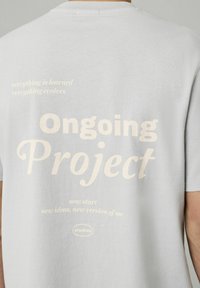 T-shirt gris clair avec un grand texte beige doux au dos : « Projet en cours » et plusieurs phrases motivantes dans des polices variées.