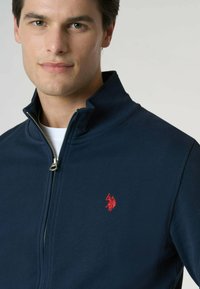 Giacca navy con zip e colletto alto, realizzata in tessuto liscio, con ricamo del logo rosso sul lato sinistro del petto. La cerniera è metallica.