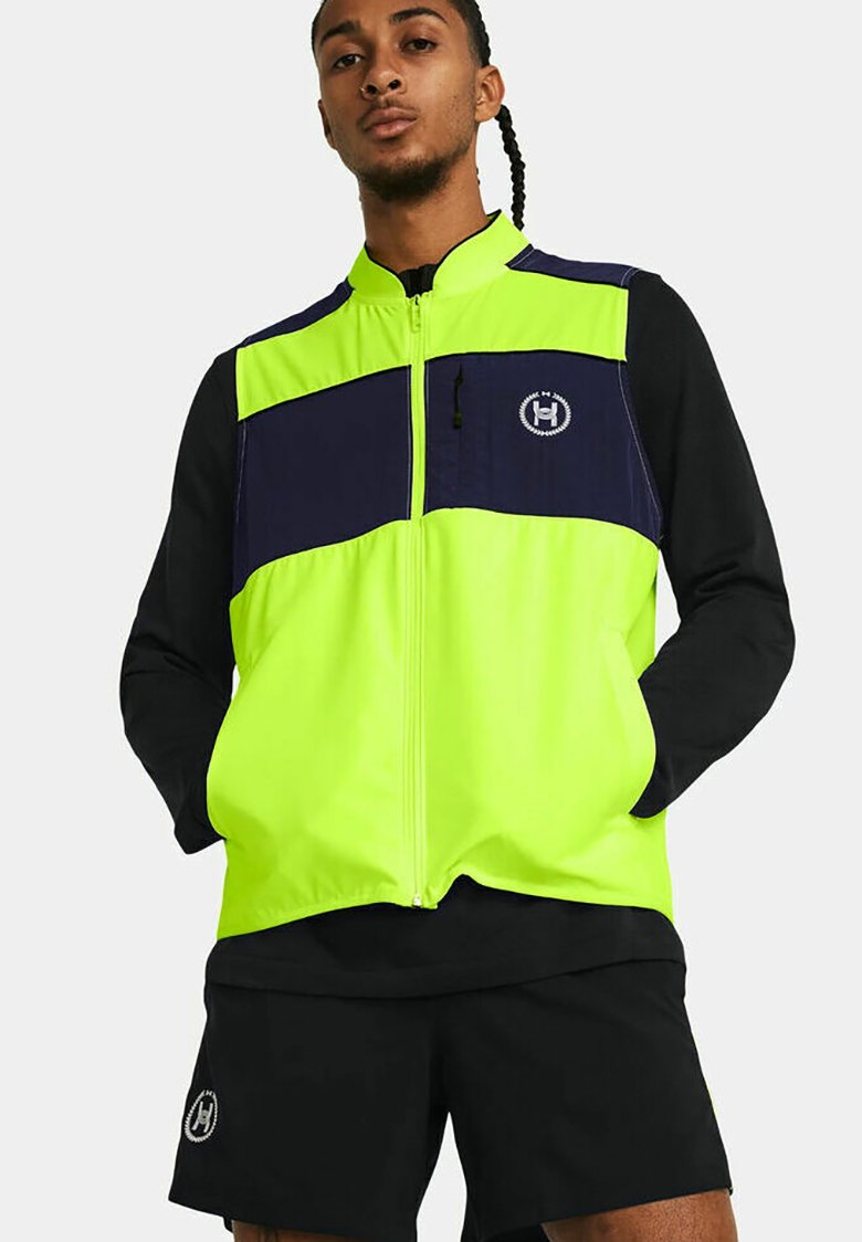 Chaqueta con cremallera en amarillo neón y marino, con mangas largas negras; cuenta con un bolsillo en el pecho y un acento de logo. Combinada con pantalones cortos negros.