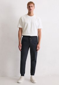 Weißes T-Shirt mit kurzen Ärmeln, kombiniert mit dunkelgrauen Jogginghosen mit elastischem Bund und Bündchen, ergänzt durch helle Sneakers.