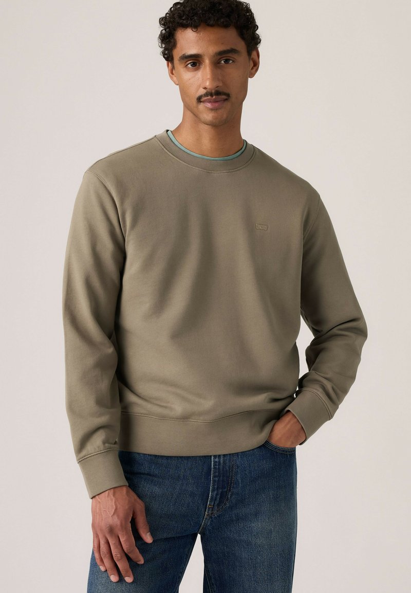 Mann mit lockigem Haar und Schnurrbart trägt olivgrünes Sweatshirt über einem hellgrünen Hemd und blauen Jeans, steht mit einer Hand in der Tasche.