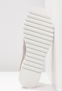 Semelle blanche striée d'une sneaker beige appuyée contre un coin blanc avec le logo "Gabor" embossé près du talon.