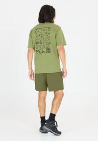 Oliivinvihreä t-paita, jossa on mustat graafiset kuviot selässä; yhdistettyinä samansävyisiin oliivinvihreisiin shortseihin. Mallilla on päällään mustat vaelluskenkä ja nilkkasukat.