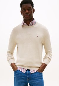 Homme noir portant un pull beige par-dessus une chemise rayée rouge et blanche, un jean bleu, se tenant avec les mains dans les poches devant un fond blanc.