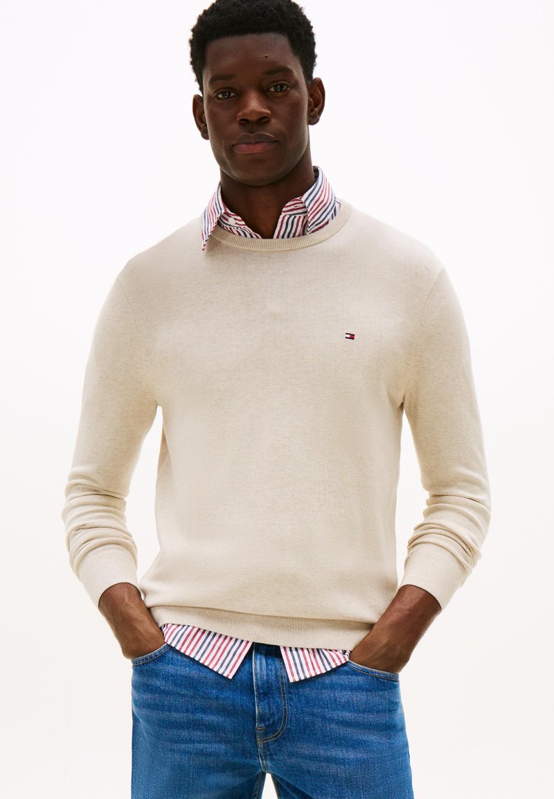 Homme noir portant un pull beige par-dessus une chemise rayée rouge et blanche, un jean bleu, se tenant avec les mains dans les poches devant un fond blanc.