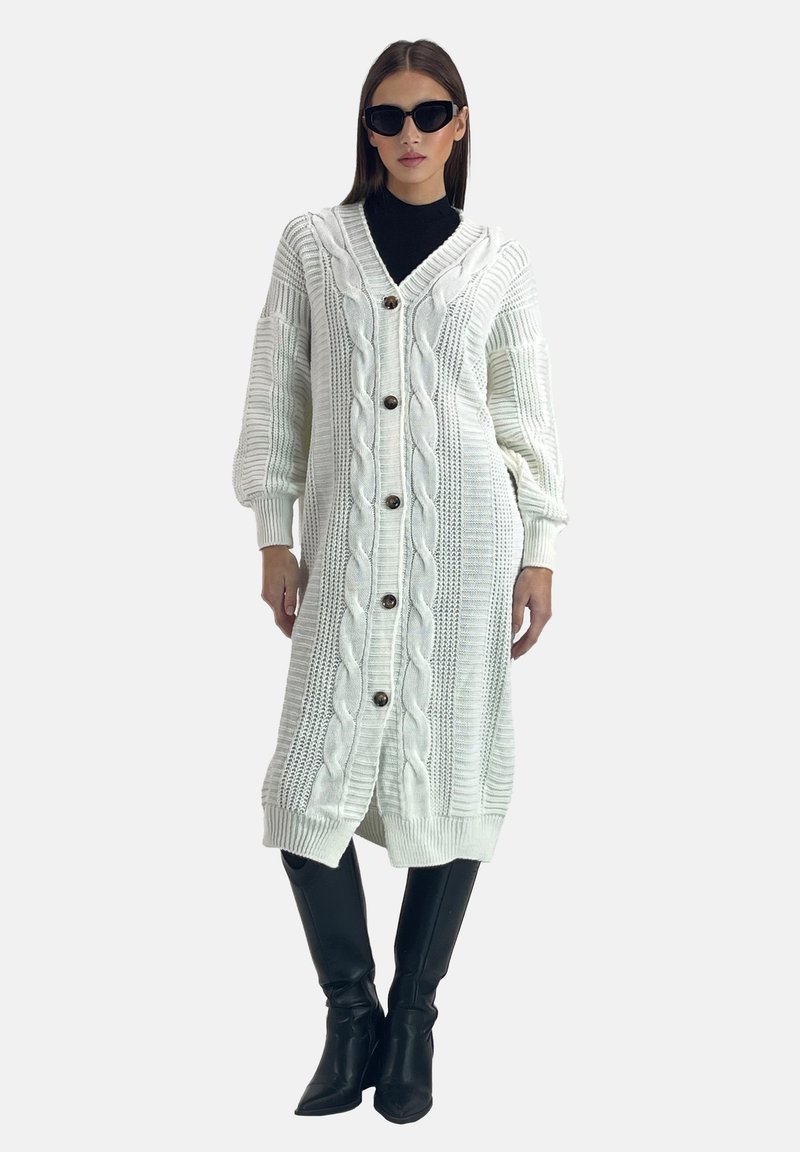Lang hvid cardigan lavet af struktureret strik med kabelmønstre, store knapper foran og ribbede manchetter og kant. Båret over en sort top og bukser.