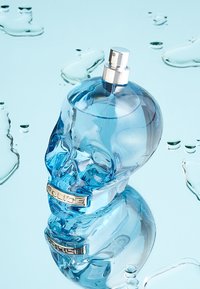 Flacon de parfum en verre bleu en forme de tête de mort avec un pulvérisateur silver et une étiquette métallique indiquant "POLICE". Réfléchi sur une surface aquatique.