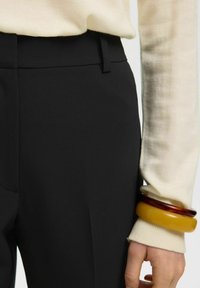 Gros plan d'une personne portant un pantalon noir et un pull crème, avec deux bracelets épais au poignet, l'un ambre et l'autre jaune.