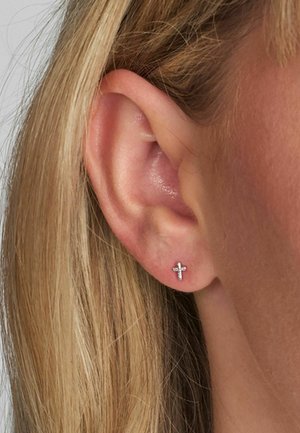 Malé strieborné náušnice v tvare kríža, s lesklým povrchom a jemným dizajnom, nosené na ušnom laloku pri svetlo blond vlasoch.