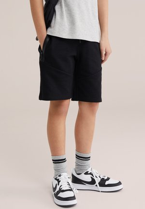 Persoon draagt zwarte korte broek, grijs t-shirt, grijze sokken met zwarte strepen en witte en zwarte Nike-sneakers, staand met handen in de zakken.