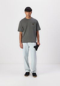 Gray oversized T-shirt with a chest pocket and yellow logo, paired with light blue jeans and black sneakers. Simple, casual fit.