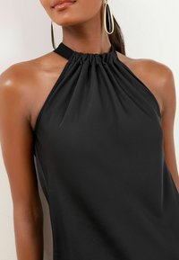 Melnā halter top ar savāktu apakšmalu un mīkstu, gludu audumu. Piedāvā brīvu piegriezumu, izceļot kailos plecus un tīru siluetu.