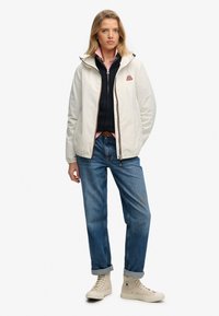 Lekka kremowa kurtka z zamkiem, kapturem i haftowanym logo. Nosi się ją na granatowym swetrze i różowej koszuli, w zestawieniu z niebieskimi dżinsami i beżowymi trampkami.