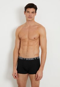 Fekete boxer alsónadrág fehér márkázott derékszíjjal, amelyen a "GUESS" felirat olvasható. Az anyag simának és illeszkedőnek tűnik. Nincsenek további minták.