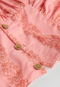 Rosa tyg med blommönster i orange nyanser. Har träknappar och drapering längs halslinjen. Mjuk, lätt textur.