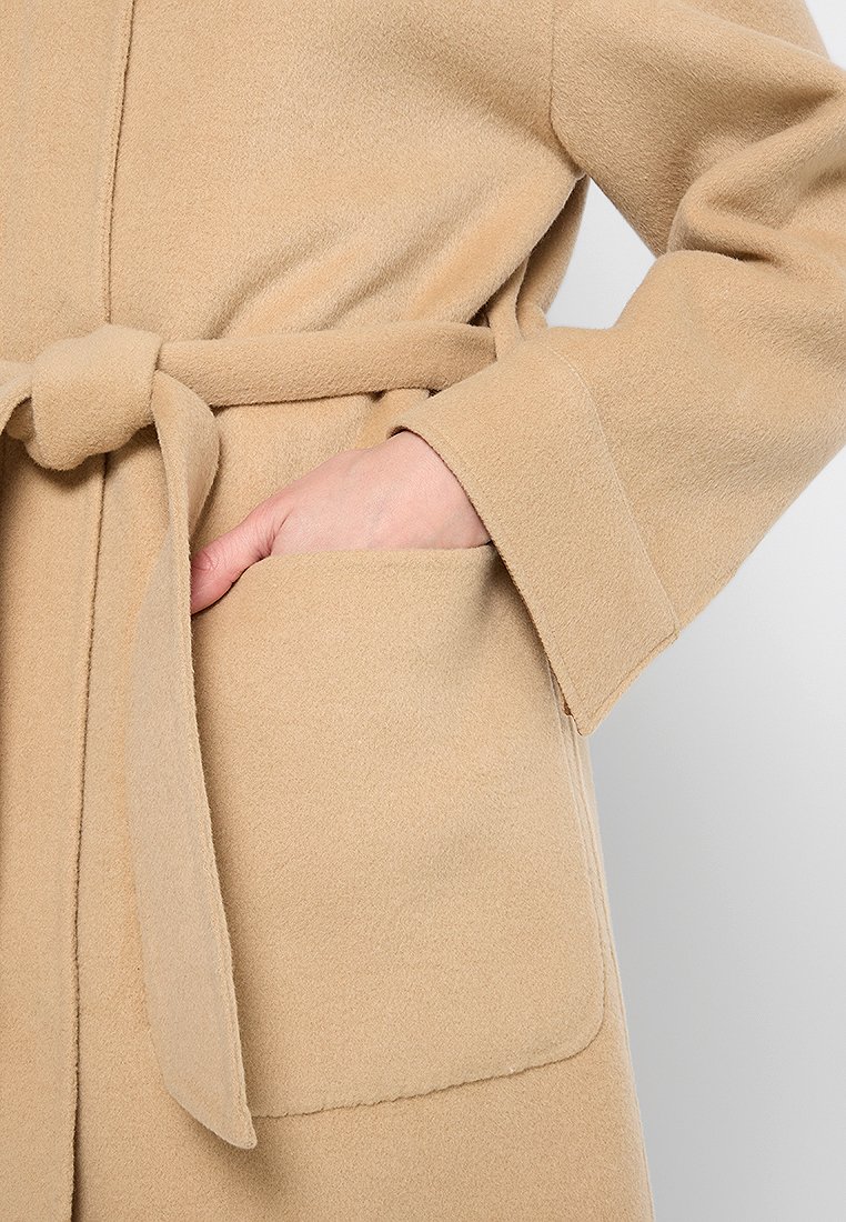 Abrigo de lana beige con un cinturón atado a la cintura, mangas anchas y un bolsillo frontal. La textura parece suave y ligeramente cepillada.